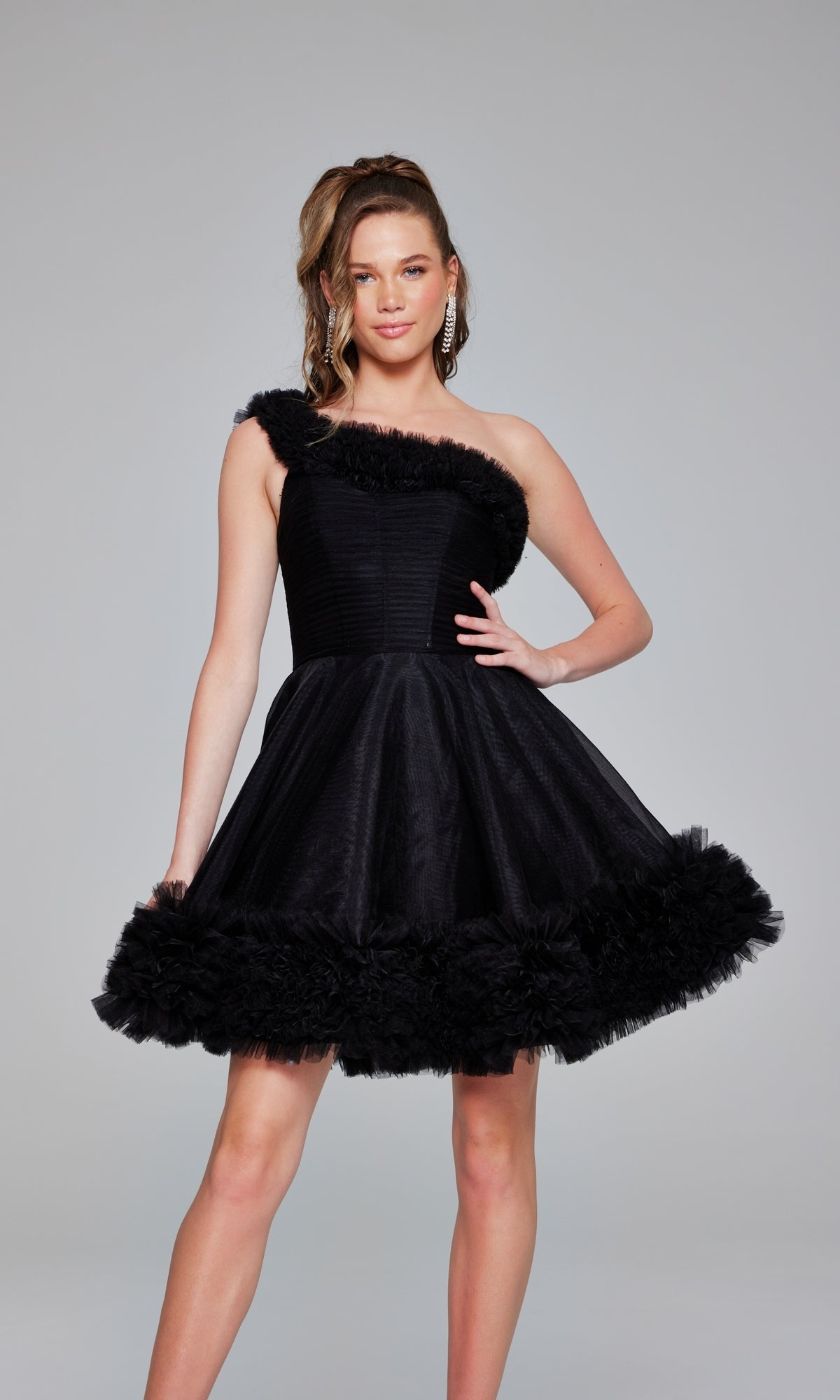 A woman poses in a studio wearing the Jovani 39665 One-Shoulder Short Ruffle Hoco Dress, a black dress featuring a ruffled neckline and hem, with one hand on her hip.